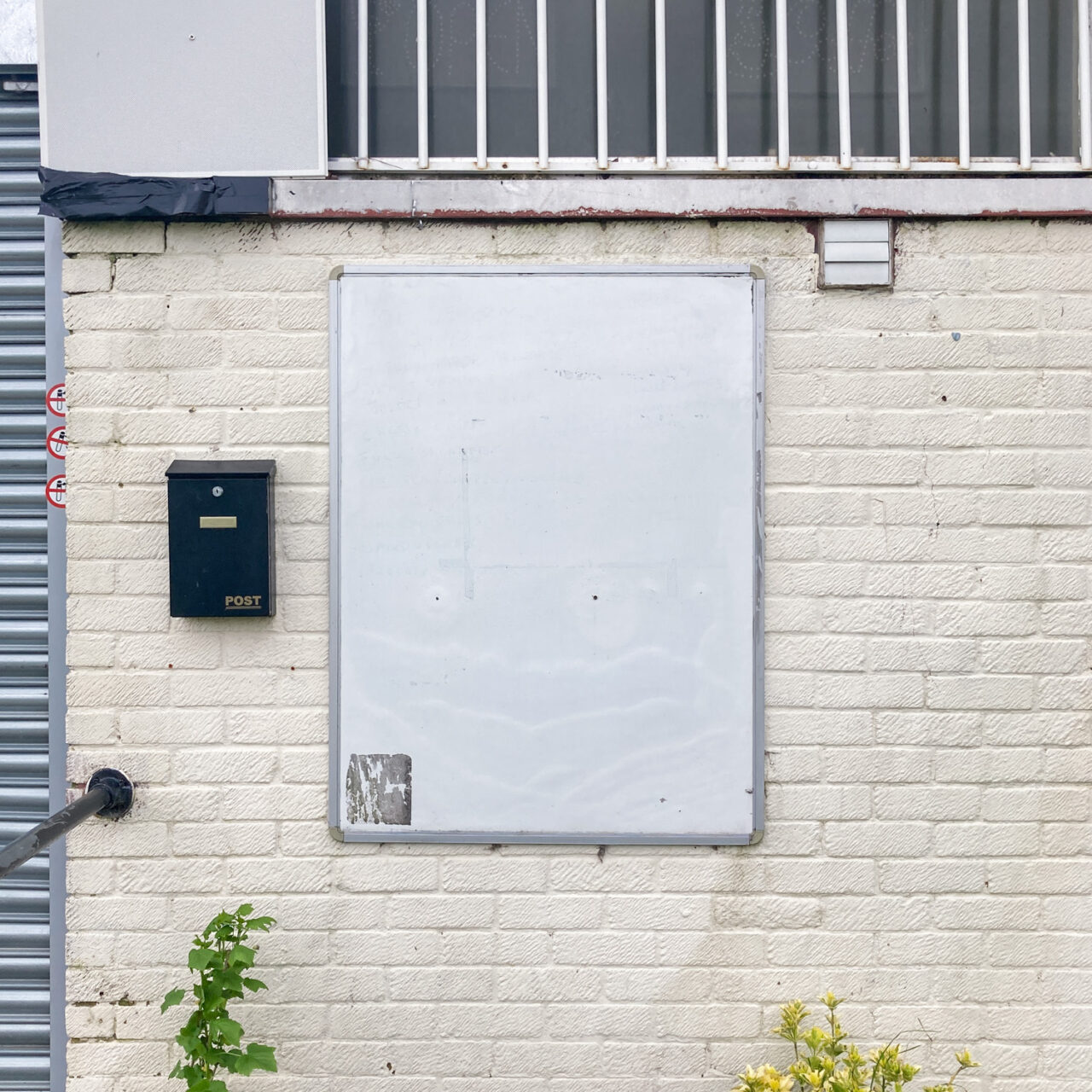 Photograph of a blank white whiteboard with a metal frame, mounted on a white-painted brick wall, with a black post box to the left, with a slither of metal shutter visible to the left and little bits of plant at the bottom of the frame.