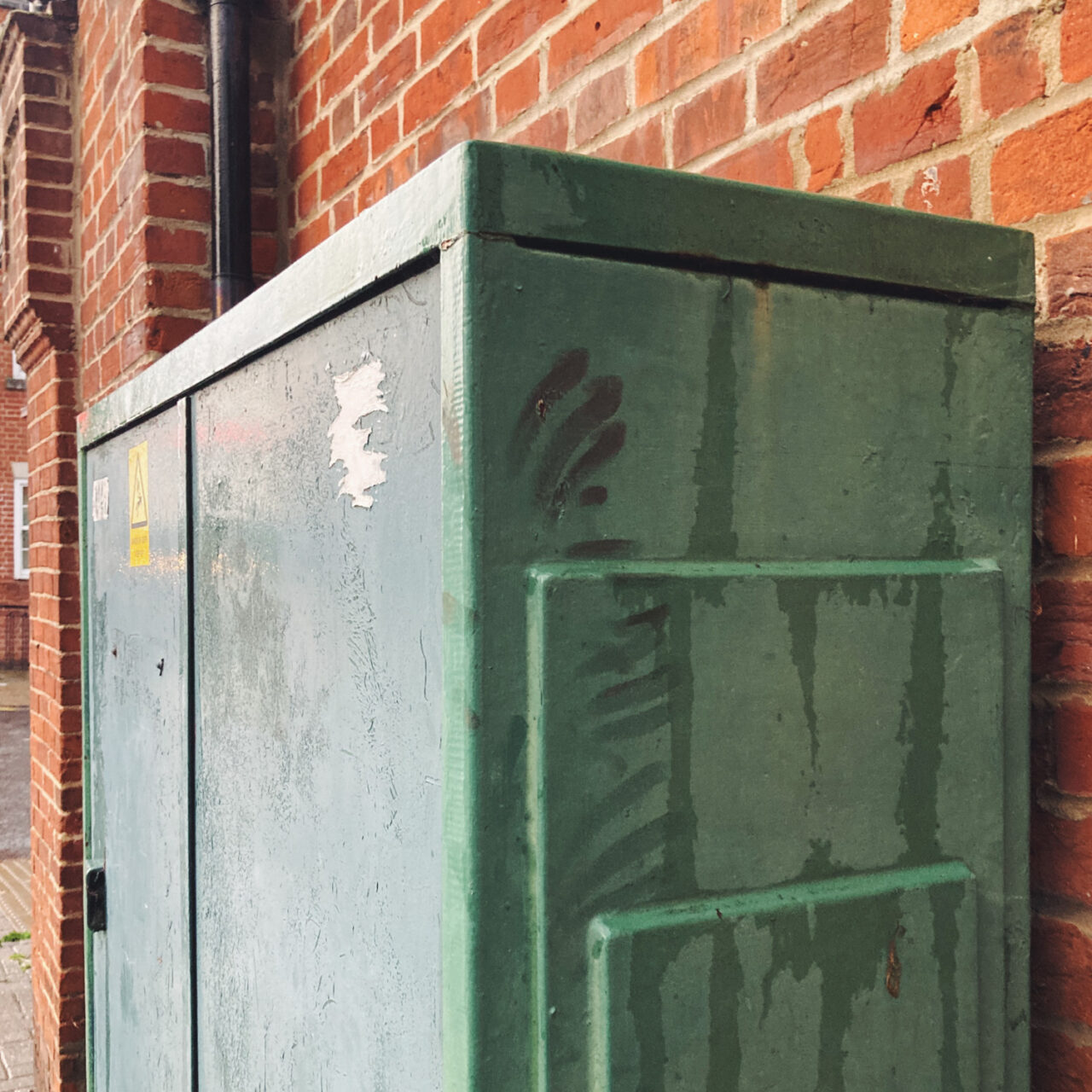 Photograph of a green telephone connection box in front of a brick walled-building, with greasy hand smears on the side.