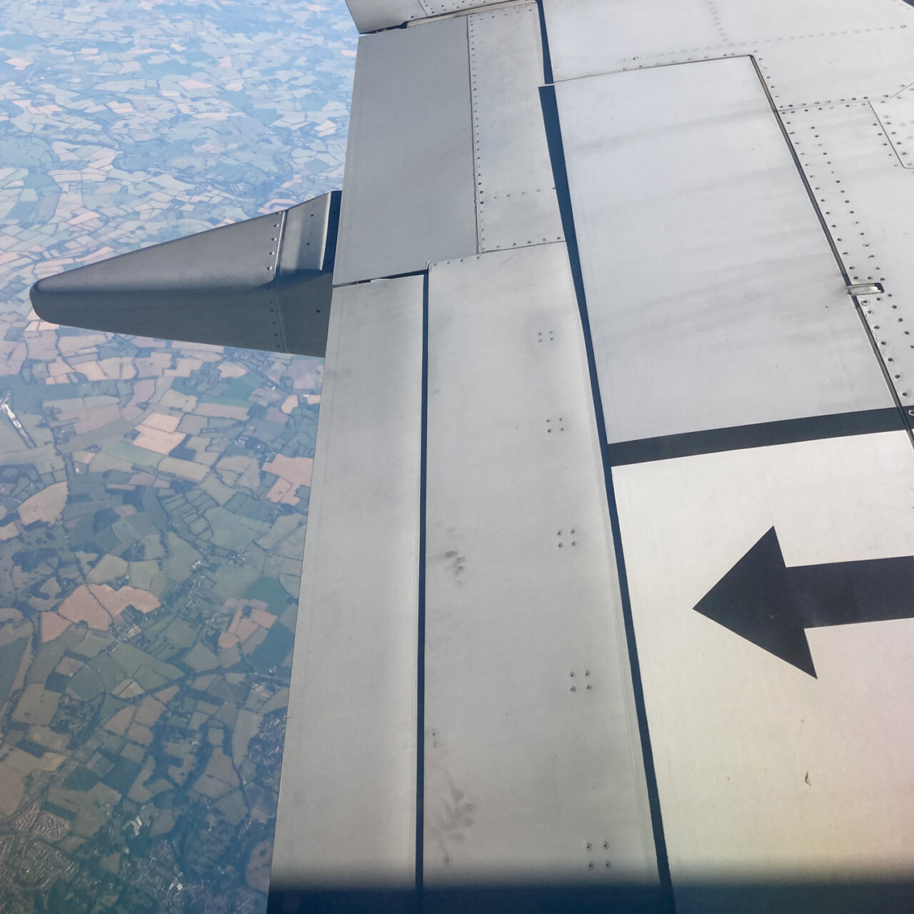 Photograph of a plane wing with dirty hand marks in the lower portion of the frame, with a black arrow to the right pointing left, fields are visible in the distance behind, to the left of frame.