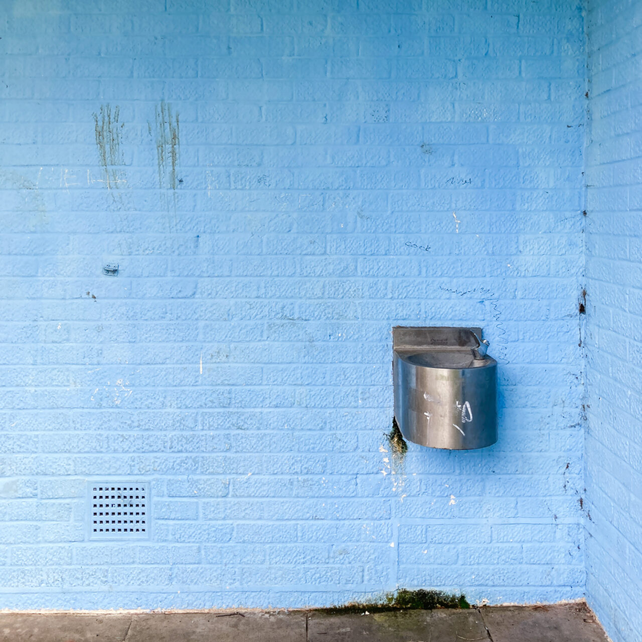 Photograph of a blue painted brick wall with two brown handprints smeared to the left and a metal drinking fountain to the lower right.