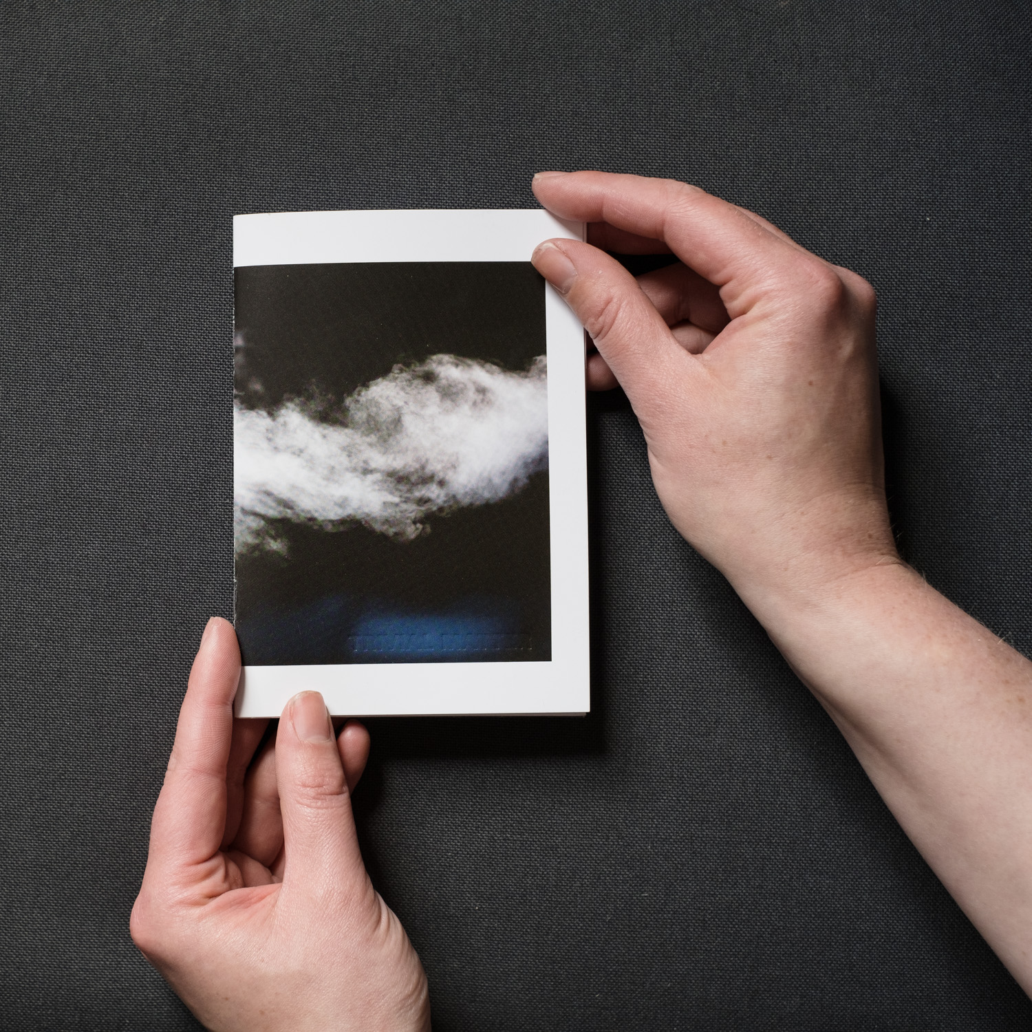 TRIVIAL MATTER 01 Photograph of TRIVIAL MATTER 01, a photo zine, on a dark grey fabric background, held by hands. Shown is the front cover with a photograph of steam on a dark background.