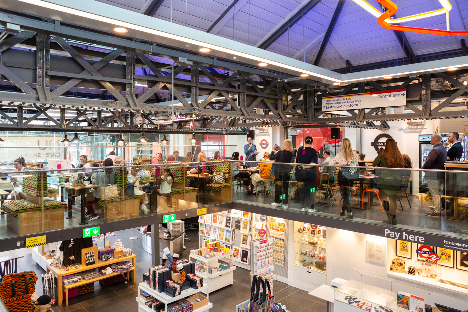 Kids in Museums Family Café Standard Launch at the London Transport Museum. Photograph by Jayne Lloyd for Kids in Museums. Photograph of the Kids in Museums Family Café Standard Launch, showing the café from a distance, with the shop below, filled with products and displays, the café filled with people with seating in booths on the left and tables and chairs to the right, visible through a glass wall. People are listening to a speech from Vanessa Eke, in the centre holding a microphone, and there is London Underground signage and strip lighting above.