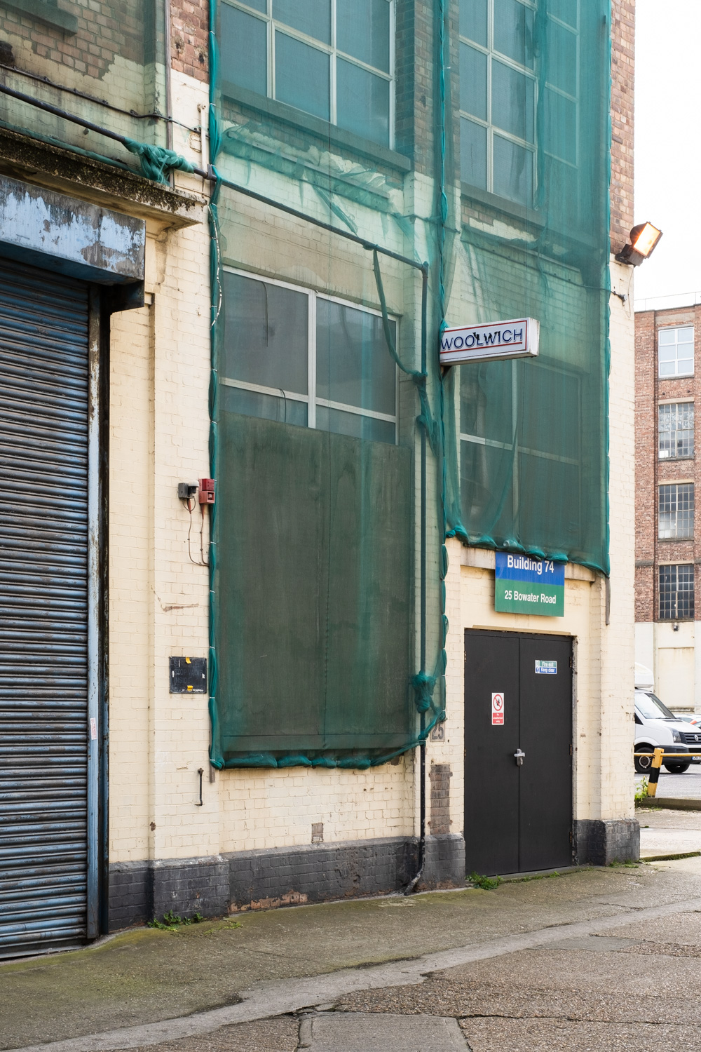 Photograph of an industrial building with green netting across the windows, a double door to the bottom right, a closed shutter to the left and an old 'WOOLWICH' sign poking out slightly above the centre.