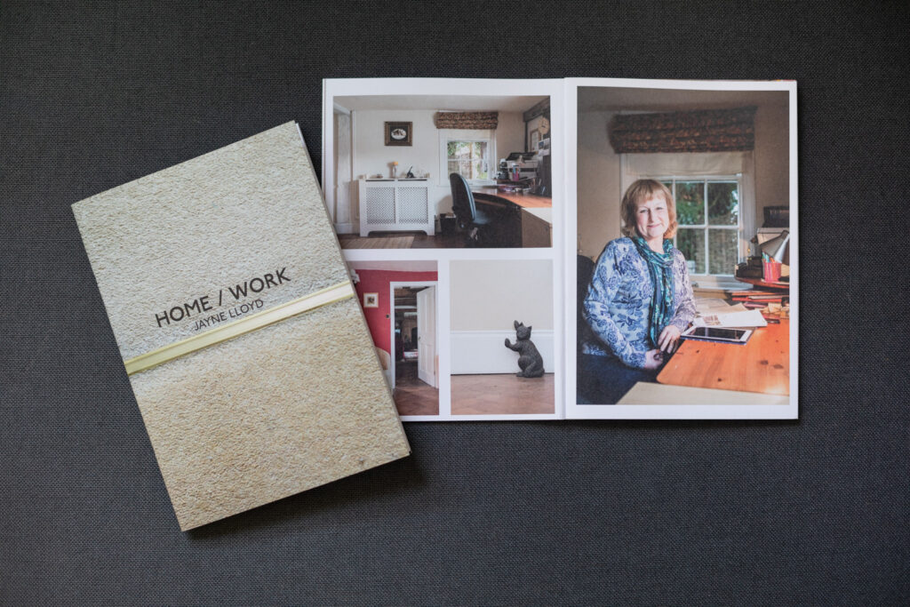 Photograph of two copies of an A6 zine on a dark grey fabric background. The copy to the left is at an angle, closed to show the front cover, a full-bleed photograph of a carpet with a metal divider, lighter at the top than at the bottom, with 'HOME / WORK, JAYNE LLOYD' in black text in the centre. Behind and to the right is the zine open on a spread showing photographs in a home office, a cat doorstop and a portrait of a woman sitting at a desk, smiling, to the right.