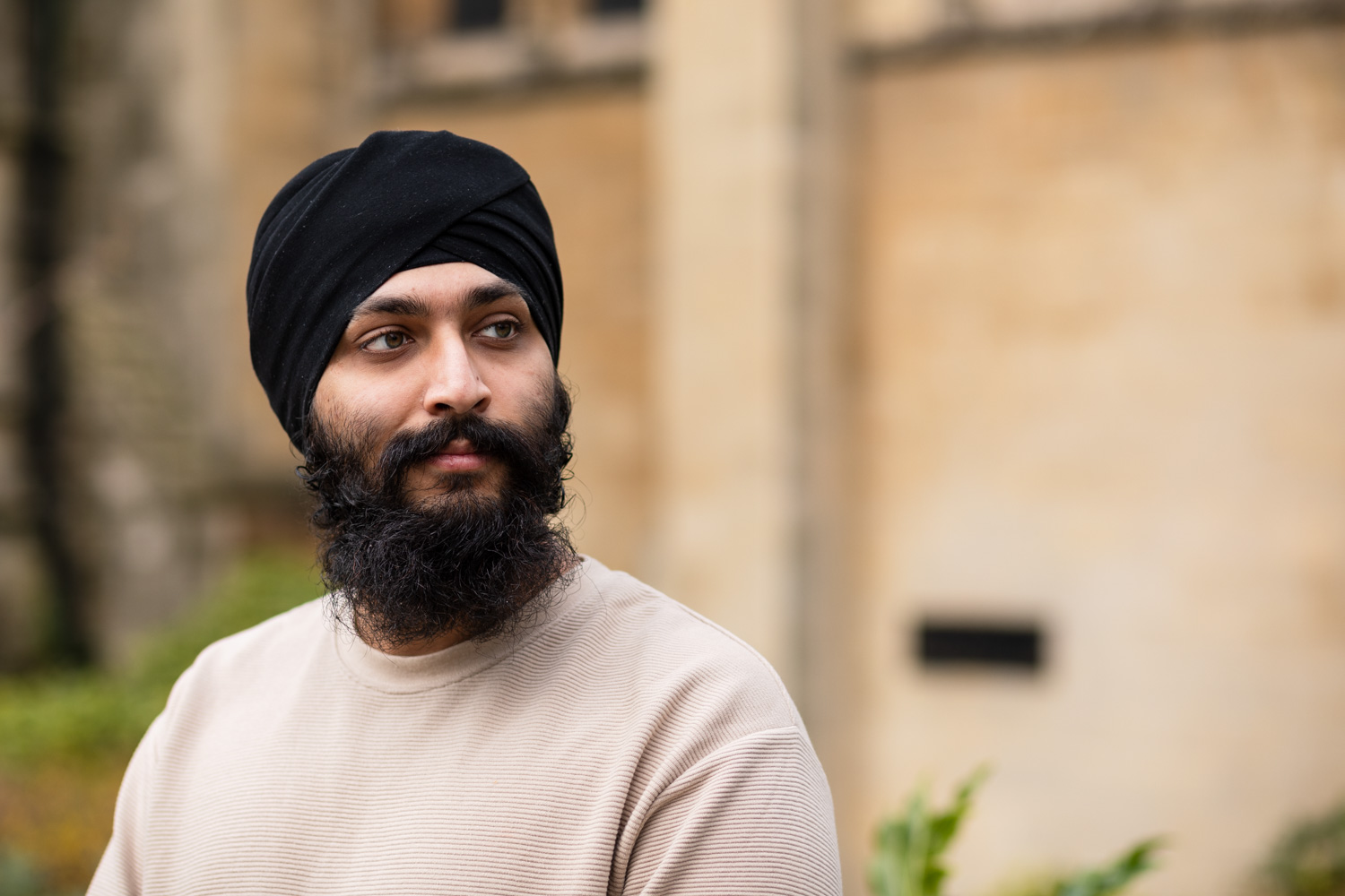 Photograph of a man with a beard wearing a black turban and beige sweatshirt, looking to the right side of the frame. He is positioned to the left and behind him a sandy-coloured building and bits of folliage are out of focus.