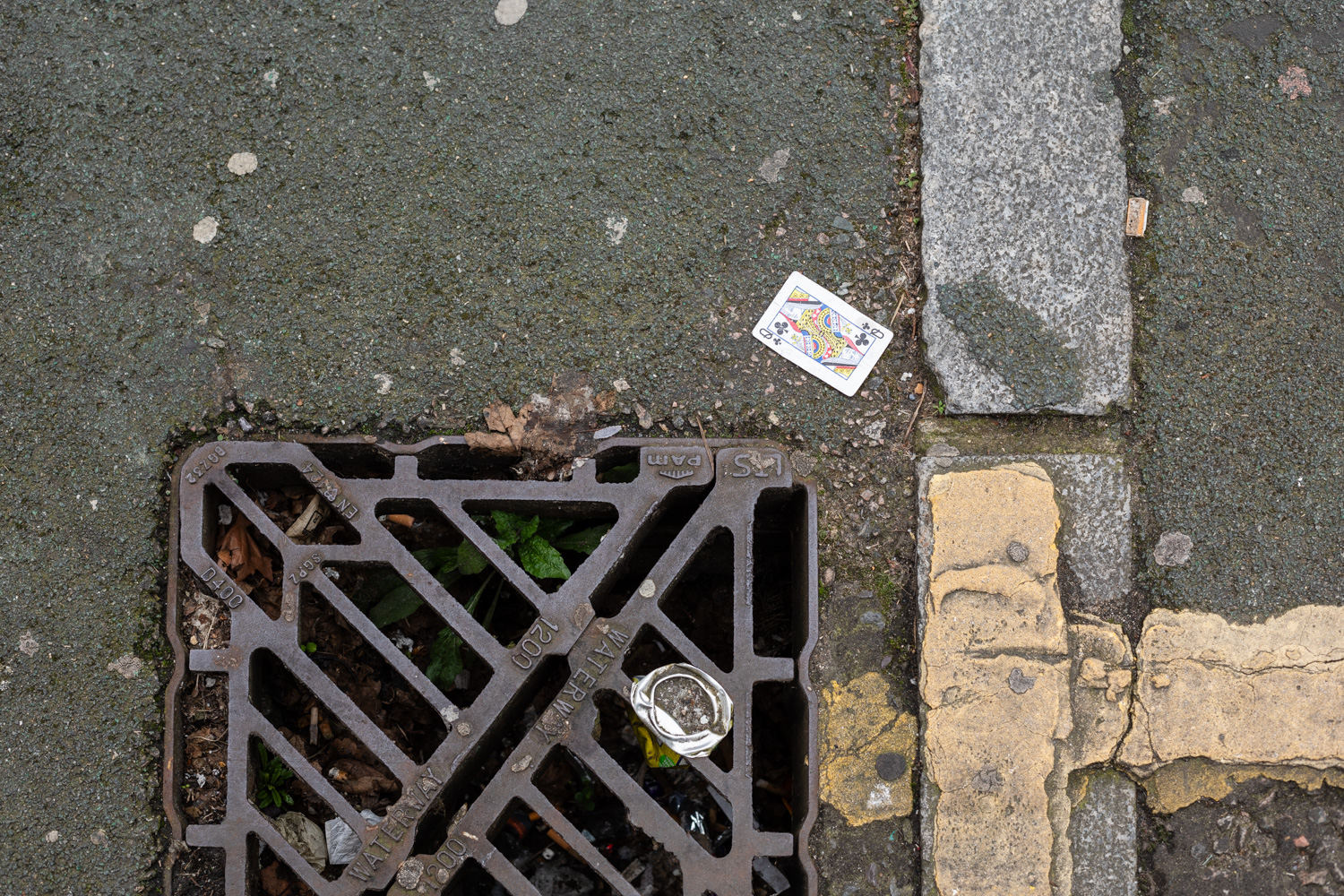 Photograph looking down at a Queen of spades playing card on the floor, with a drain at the bottom of the frame, and paving and paint elements to the right.