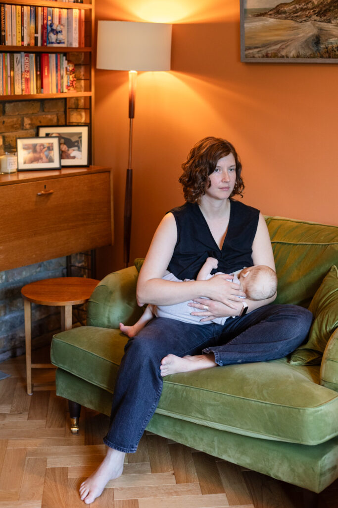 Photograph of a woman in a black top and dark jeans, breastfeeding a baby whilst sat on a green velvet sofa, looking towards the right of the frame. Behind her a standard lamp throws warm light onto the terracotta wall, with an oil painting above and to the righ, a wooden dresser filled with books and framed photographs to the left with a small wooden table below, and a wooden floor below.