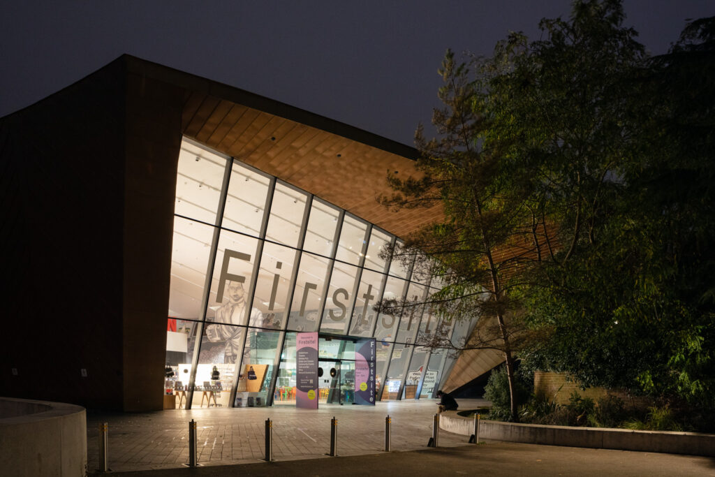 Firstsite at Night. Photograph by Jayne Lloyd. Photograph of the Firstsite gallery building at night, glowing, with a dark sky above, trees to the right and metal bollards in the foreground. Artworks, the shop and café can be seen inside, with 'Firstsite' written in large letters across the glass front.