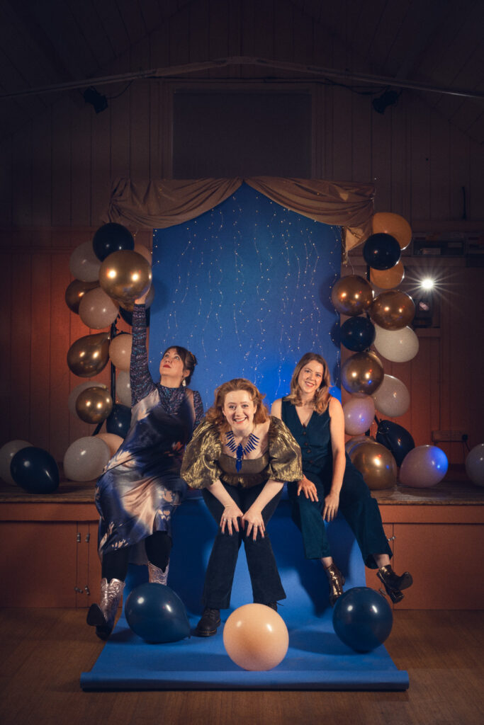 Wise Woman, photographed by Jayne Lloyd at Blaxhall Village Hall. Photograph of a stage in a traditional village hall, dark around the edges but with blue background paper on a stand lit in the centre of the frame and falling over onto the wooden floor below. The background is topped with gold fabric, framed by gold, white, peach and navy balloown, which also spill onto the floor below and has fairy tiny lights hanging across the centre and top. A light with a starburst effect shines to the right and wires and light stands are visible at the sides. Three women dressed in blues and golds sit on the edge of the stage, playing with the balloons and smiling and laughing towards the camera.