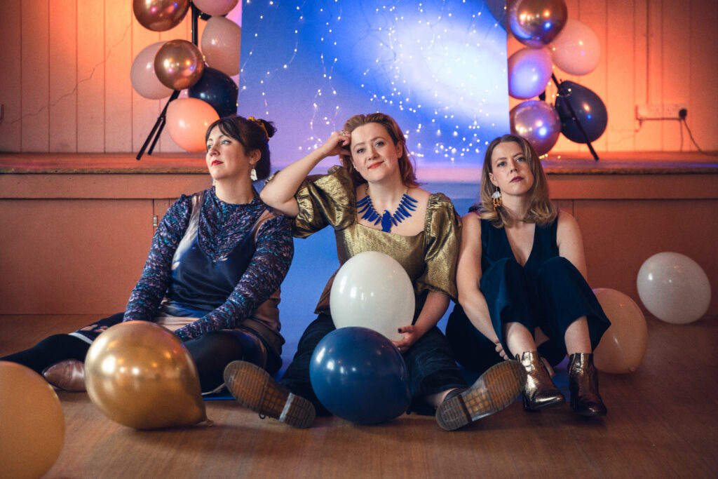 Wise Woman, photographed by Jayne Lloyd at Blaxhall Village Hall. Photograph of a three women sitting on a wooden floor in front of a low stage in a village hall, with gold, white, navy and peach balloons around them. Behind them, on the stage, a blue background on a stand is decorated with tiny fairy lights and balloons either side, with spot lights highlighting some areas.