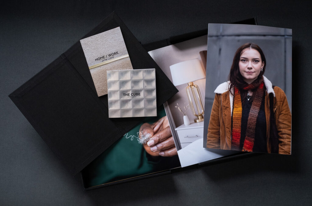 Photograph of a black box of prints on a dark grey background, with three prints visible showing a hand holding bent silver wire, an interior with a lamp and vase on a bedside cabinet and a portrait of a woman in a brown coat and scarf. There's a smaller black folder on top with two small publications on top, titled 'HOME / WORK' and 'THE CUBE'